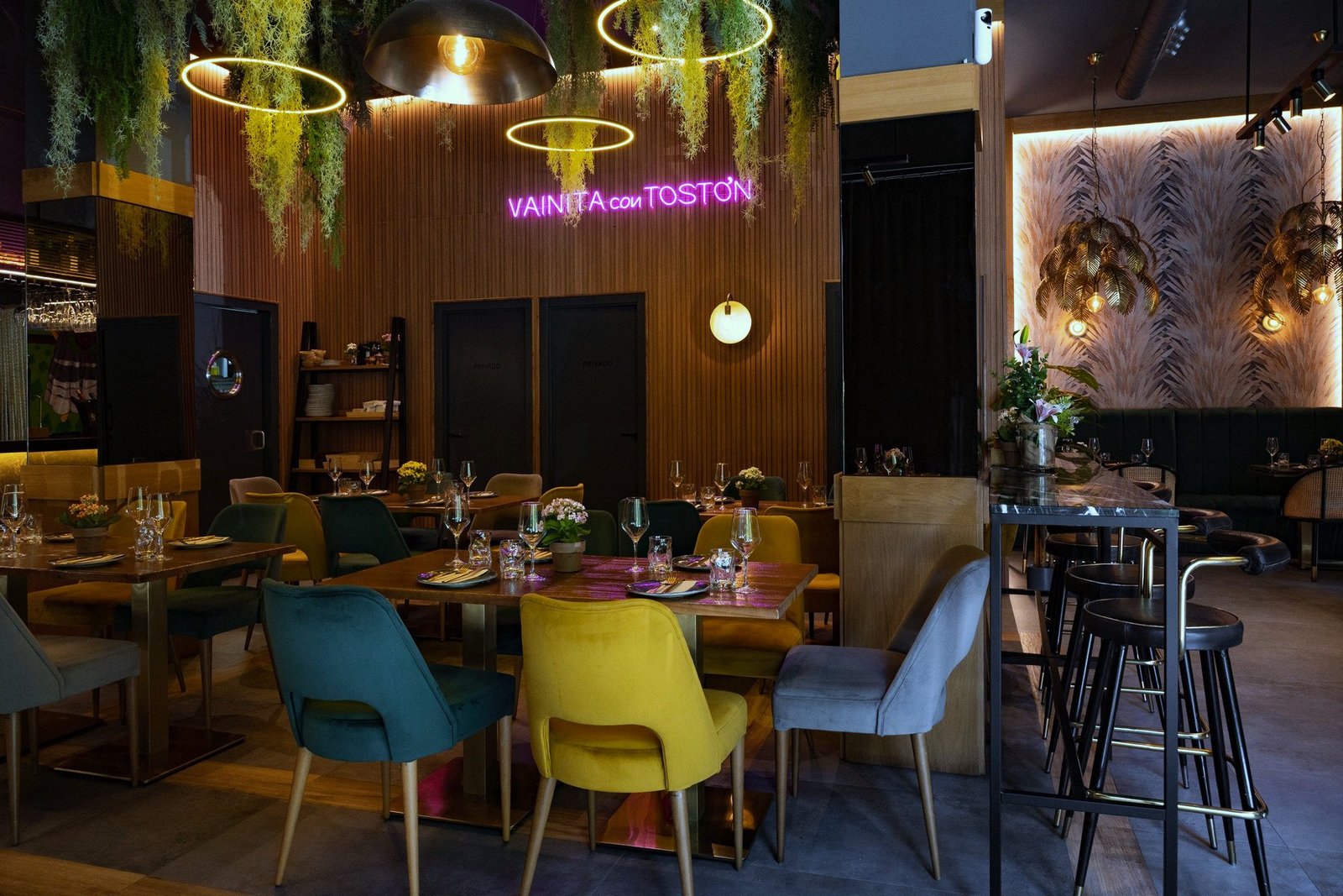 Panorámica de la sala con sillas de terciopelo de colores, plantas colgantes y el rótulo de neón